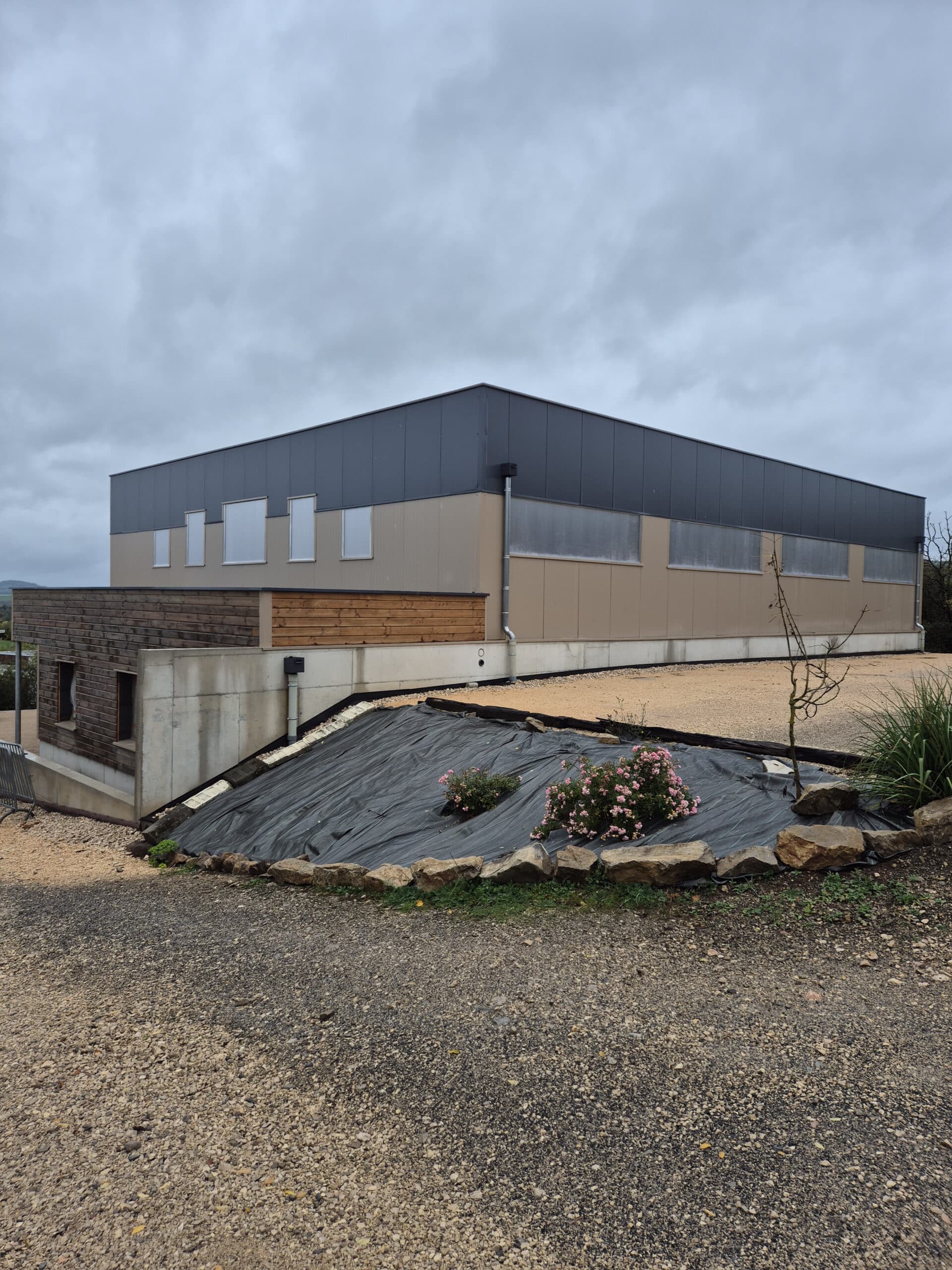 Bâtiment industriel moderne en bardage métal gris et beige. Aménagement paysager en gravier et talus protégé par géotextile avec rosiers roses.