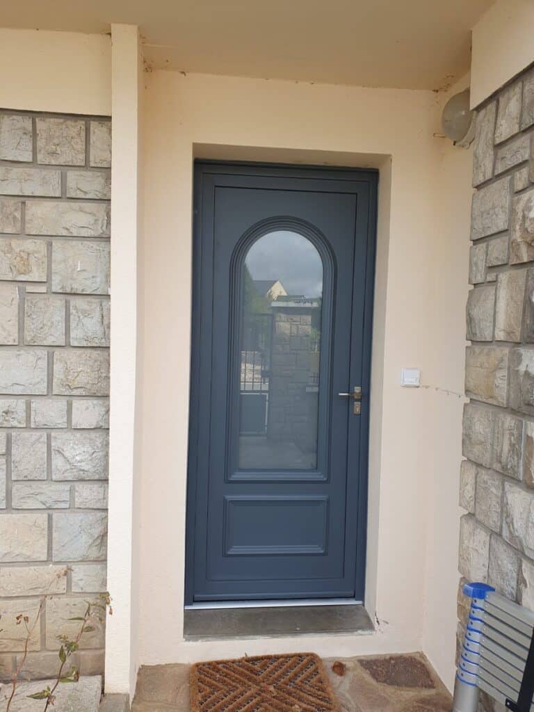 Porte d'entrée gris anthracite avec vitrage cintré Porte d'entrée gris anthracite avec vitrage cintré, sur une façade en crépi beige et pierre apparente.